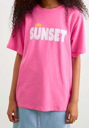 Personne portant un t-shirt rose vif avec le mot "SUNSET" et un graphique de soleil jaune au-dessus de la lettre U, associé à une jupe en jean bleu clair.
