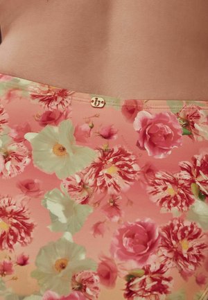 Prim-plan al unui material roz cu model floral cu flori roșii, albe și verzi purtat în talie, arătând o mică etichetă aurie de marcă.