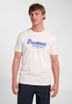 Mann mit Schnurrbart trägt weißes hummel T-Shirt mit blauem Logo und Text, steht vor schlichten hellem Hintergrund.