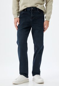 Vaqueros de mezclilla azul oscuro con corte recto, que cuentan con cierre de botón y cremallera, bolsillos frontales y una textura suave. Combinados con zapatillas blancas.