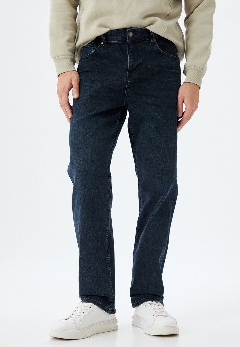 Vaqueros de mezclilla azul oscuro con corte recto, que cuentan con cierre de botón y cremallera, bolsillos frontales y una textura suave. Combinados con zapatillas blancas.