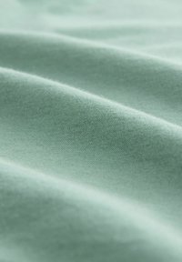 Gaiši zaļš kokvilnas audums ar maigu tekstūru, ar maigiem krokām un gludu virsmu, demonstrējot vienveidīgu krāsu bez rakstiem.