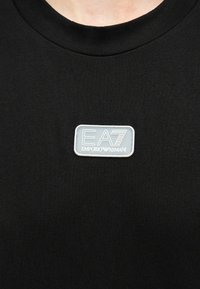 Μαύρο μπλουζάκι με στρογγυλή λαιμόκοψη και ένα μικρό λευκό ορθογώνιο ταμπελάκι με την επιγραφή "EA7 Emporio Armani" στο κέντρο του στήθους.