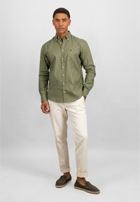 Groene button-up shirt met een zak, beige broek en donkergroene instapschoenen. Glad stof, getailleerde pasvorm en opgerolde broekspijpen.
