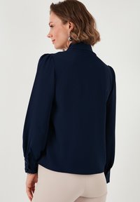 Marineblauwe blouse met lange, opgeblazen mouwen en een hoge kraag. Gladde stof en knoopdetails bij de manchetten. Eenvoudig, elegant ontwerp.