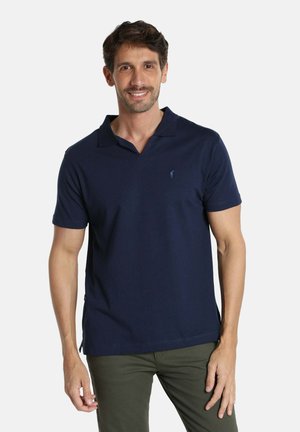 Homme portant un polo bleu marine à manches courtes et un pantalon vert olive, souriant, debout devant un fond blanc uni.