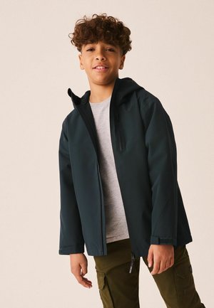 Jongen met krullend haar die een donker capuchonjack draagt over een grijs shirt en olijfgroene cargobroek, staand tegen een effen achtergrond.