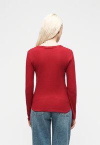 Γυναίκα με μακριά ξανθά μαλλιά, φορόντας εφαρμοστό κόκκινο μπλουζάκι μακρυμάνικο και μπλε τζιν, όρθια με την πλάτη στραμμένη σε απλό φόντο.