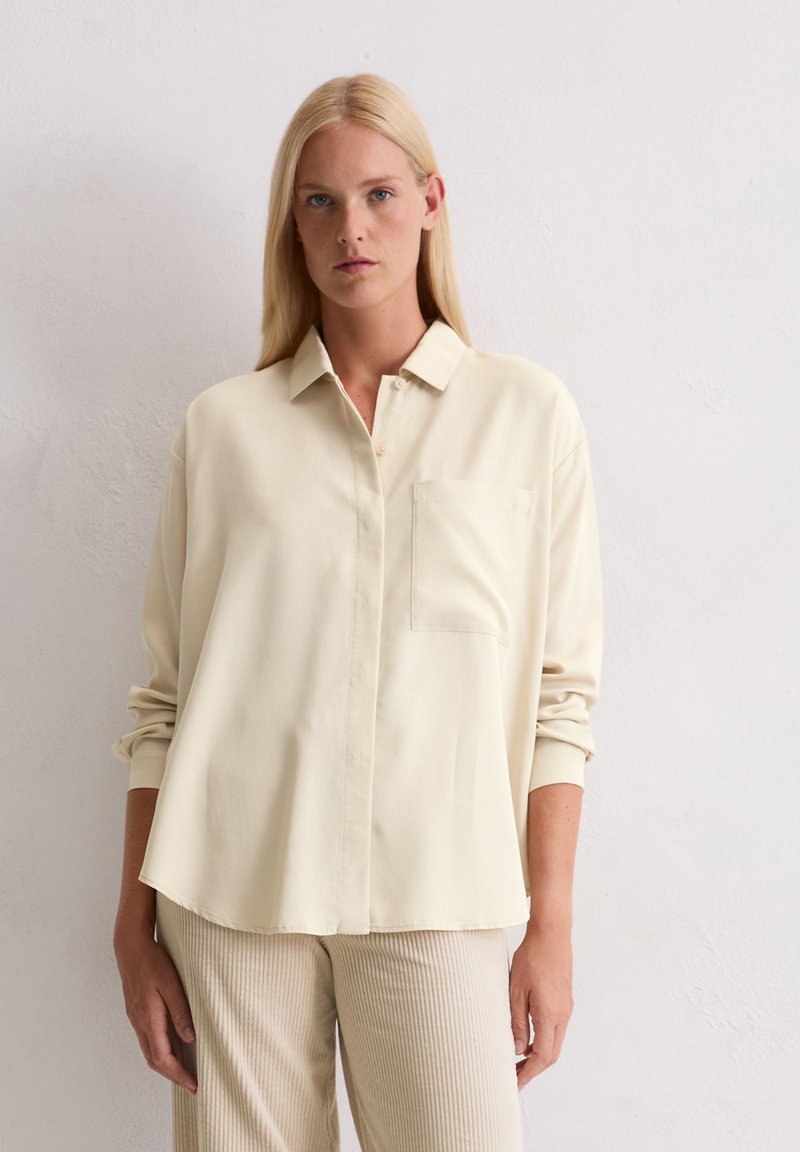 Lichtbeige blouse met knopen, een kraag, lange mouwen en een borstzak; glad materiaal met een relaxte pasvorm. Eenvoudig ontwerp.