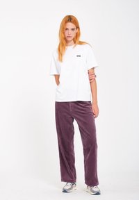 Camisa de algodón blanca con un pequeño logo negro en el pecho, combinada con pantalones de pana anchos en color morado y zapatillas grises con acentos en blanco.