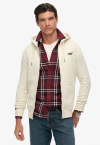 Cremefarvet zip-up hoodie med snøre i hætten, lagdelt over en rødternet skjorte, parret med blå jeans. Har detaljeret syning og et logo.