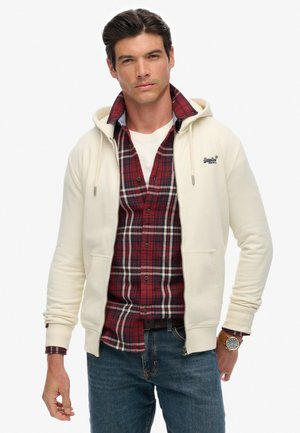 Un hoodie crème zippé avec une capuche à cordon, superposé sur une chemise à carreaux rouges, associé à un jean bleu. Présente des coutures détaillées et un logo.
