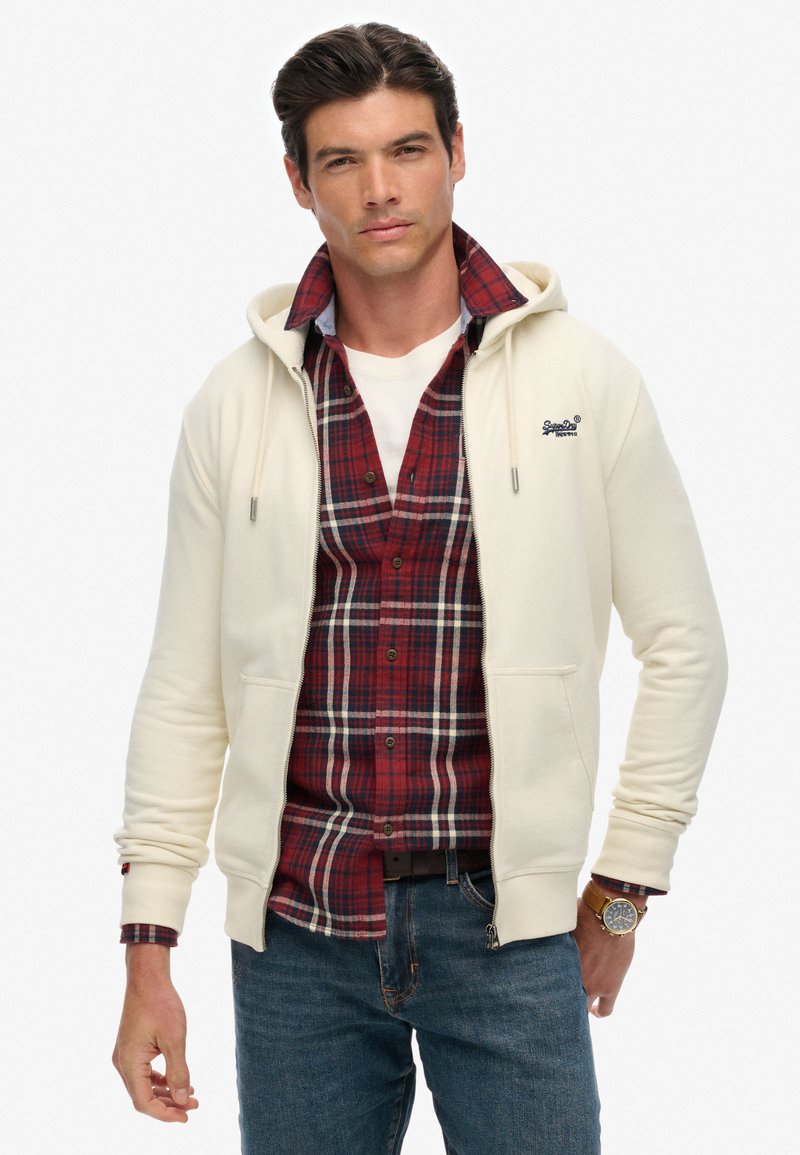 Cremefarbener Zip-Hoodie mit Kordelzugkapuze, über einem roten Karoshemd getragen, kombiniert mit blauen Jeans. Verfügt über detaillierte Nähte und ein Logo.