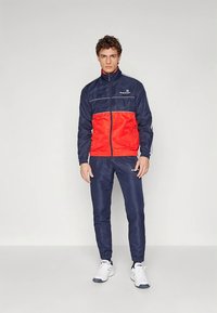 Sporta tērps tumsnējs zilā un sarkanā krāsā; žakete ar rāvējslēdzēju, augšējā daļa tumsnējs zila, apakšējā sarkana, sudraba akcenti, atbilstošas bikses, gluds materiāls un elastīgas aproces.