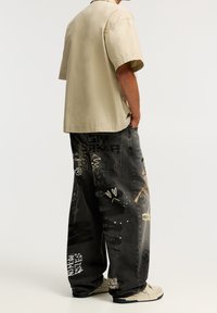 Ljusbeige kortärmad skjorta kombinerad med mörkgrå lösa jeans som har olika graffitistil utskrifter och stänkmönster.