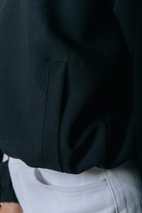 Chaqueta de tela negra con una textura sutil, costura lateral y un detalle en los puños. Combinada con pantalones de tela blanca y texturizada.