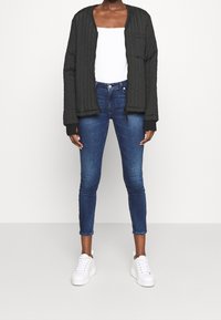 Quiltad svart bomberjacka med dragkedja och sidofickor över en vit topp, kombinerad med blå smala jeans och vita sneakers.