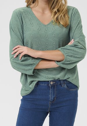 Kobieta ze skrzyżowanymi ramionami, ubrana w jasnozieloną haftowaną bluzkę z długim rękawem i niebieskie jeansy, na neutralnym tle.