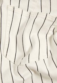 Vit tyg med vertikala svarta ränder, med en fyrkantig ficka med synliga söm detaljer. Slät textur, avslappnad design.