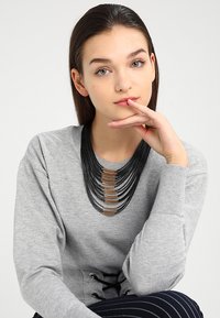 Collar de múltiples capas con hilos negros y acentos metálicos dorados. Modelo vistiendo una blusa gris con laterales con cordones, sentada con una expresión neutral.