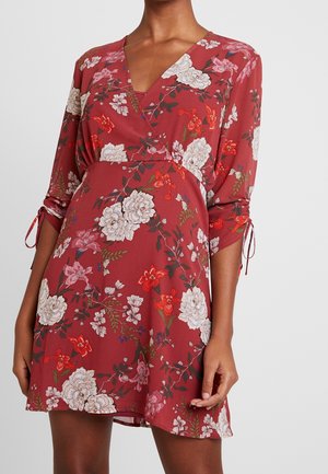 Femme portant une robe portefeuille rouge à motifs floraux, manches trois-quarts nouées aux poignets, avec des fleurs blanches, roses et rouges.