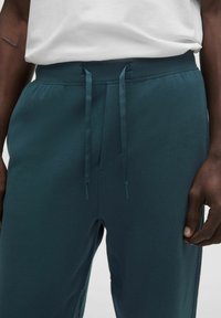 Pantalones de chándal en tono verde azulado con cintura ajustable, confeccionados con un tejido suave. Incluyen bolsillos laterales y un corte relajado; diseño minimalista.
