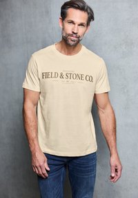 Beige tričko s krátkým rukávem s textem "FIELD & STONE CO." v tmavě hnědé barvě. Bavlněný materiál; kulatý výstřih. Nosit s modrými džínami.