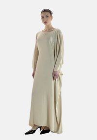 Robe beige à manches longues, longueur cheville, en tissu doux, avec un col rond et un drapé élégant sur un côté. Stylée avec des escarpins noirs.