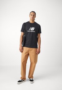 Black cotton t-shirt with large white "New Balance" logo, paired with tan straight-leg pants and black sneakers with white accents. Black bag slung over shoulder.