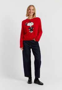 Roter Pullover mit einer Snoopy-Grafik in Schwarz und einer gepunkteten Fliege; kombiniert mit hochtaillierten dunklen Jeans und schwarzen Slippern.
