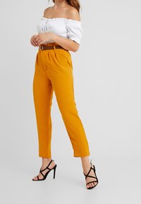 Off-the-shoulder white ribbed top, orange high-waisted trousers with pleats, and black strappy high-heeled sandals.