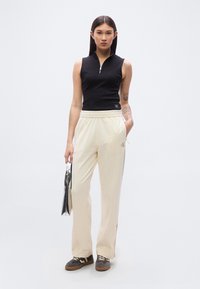 Μαύρο αμάνικο τοπ με φερμουάρ στον λαιμό, συνδυασμένο με εκρού τζόγκερ με πλαϊνές ρίγες και λογότυπο. Αξεσουάρ: μεταλλική τσάντα.