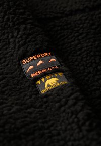 Zwart fleece stof met een gestructureerd oppervlak, met twee labels: één oranje 'SUPERDRY' en één gele 'OUTDOOR CO.' met een berenafbeelding.