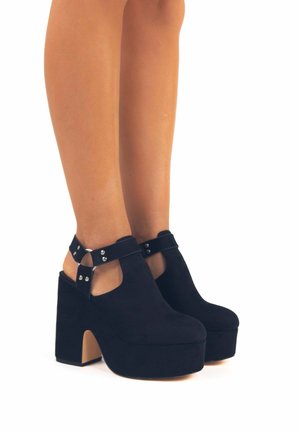 Tacchi a pianta in suede nera con un tacco spesso, schiena aperta, cinturini alla caviglia con dettagli metallici e design a punta arrotondata.