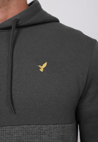 Hanorac gri închis cu o secțiune inferioară texturată și un mic colibri auriu brodat în partea superioară a pieptului din stânga.