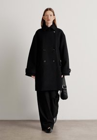 Cappotto nero doppiopetto realizzato in tessuto testurizzato con grandi bottoni, dotato di maniche ampie e tasche laterali, indossato sopra pantaloni neri larghi.