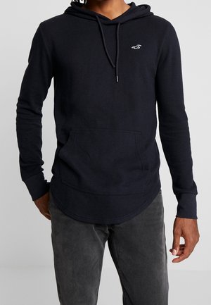 Personne portant un sweat à capuche noir avec une poche avant et un logo blanc sur la poitrine, associé à un pantalon gris foncé.