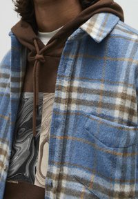 Blauw geruite flanellen overhemd met bruine en witte accenten, gemaakt van zachte stof, gelaagd over een bruine hoodie met een grafisch ontwerp.