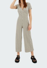 Jumpsuit rayé avec un design enveloppant, des manches courtes et un pantalon à jambes larges. Fabriqué en tissu léger dans des teintes beige et brun foncé. Sandales noires.