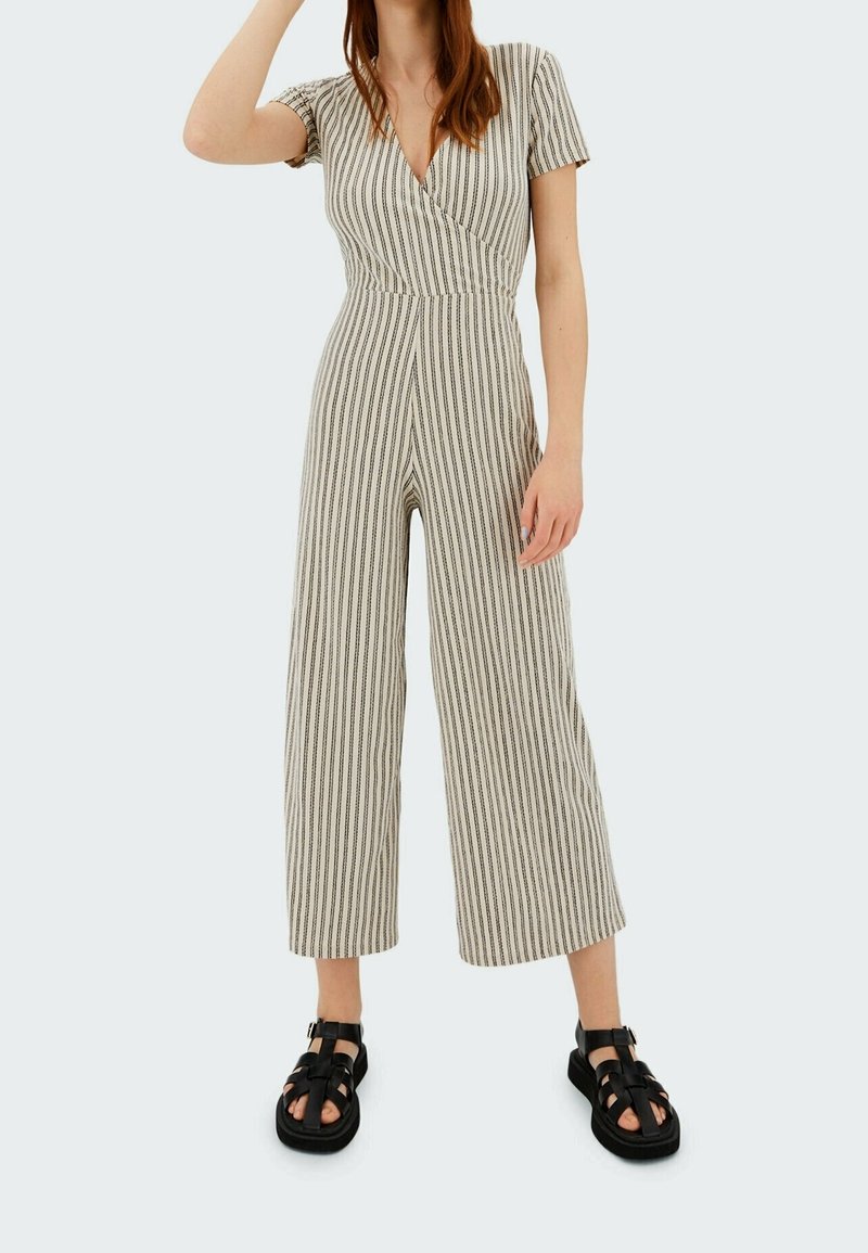 Jumpsuit rayé avec un design enveloppant, des manches courtes et un pantalon à jambes larges. Fabriqué en tissu léger dans des teintes beige et brun foncé. Sandales noires.