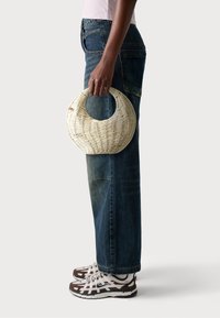 Borsa circolare intrecciata tenuta da una persona, di colore beige chiaro, superficie texture e manico. Abbinata a jeans blu scuro e scarpe da ginnastica marroni.