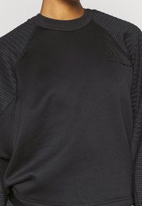 Sweat-shirt de sport noir avec texture côtelée sur les épaules, coupe ajustée et détail discret de logo sur la poitrine. Tissu doux.