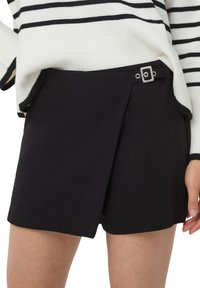 Pantaloni corti neri con piega frontale, caratterizzati da un dettaglio con fibbia quadrata in argento in vita. Il materiale è liscio e ha una vestibilità sartoriale.