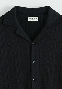 Chemise noire à manches courtes avec un motif en diamant texturé, dotée d'un col rabattu et de boutons noirs. Étiquette indiquant "FRILIVIN, taille M."