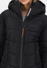 Giubbotto puffo nero con cappuccio, caratterizzato da una texture trapuntata e una zip frontale. Accentuato con un logo e un tirazip in pelle.