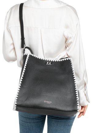 Sac à bandoulière en cuir noir texturé avec bords cousus blancs, porté en travers du corps par une personne vêtue d'un chemisier blanc et d'un jean bleu.