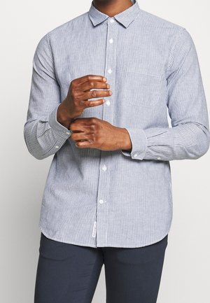 Hombre abotonando el puño de una camisa de rayas verticales azules y blancas de manga larga, vistiendo pantalones oscuros, sobre un fondo claro y liso.