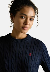 Mladá žena s kudrnatými vlasy, která má na sobě tmavě modrý pletený svetr s malým červeným výšivkou na hrudi.