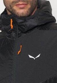 Mustmust jacket kõrge kraega, kontrastsete oranžide tõmblukkude ja logo detailidega, sile tekstuur ning geomeetriline paneeli disain.