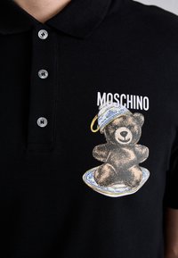 Zwarte polo met een geborduurd berenafbeelding op een decoratief bord, met de tekst "Moschino" erboven. Met knoopsluiting bij de kraag en een gestructureerde stof.